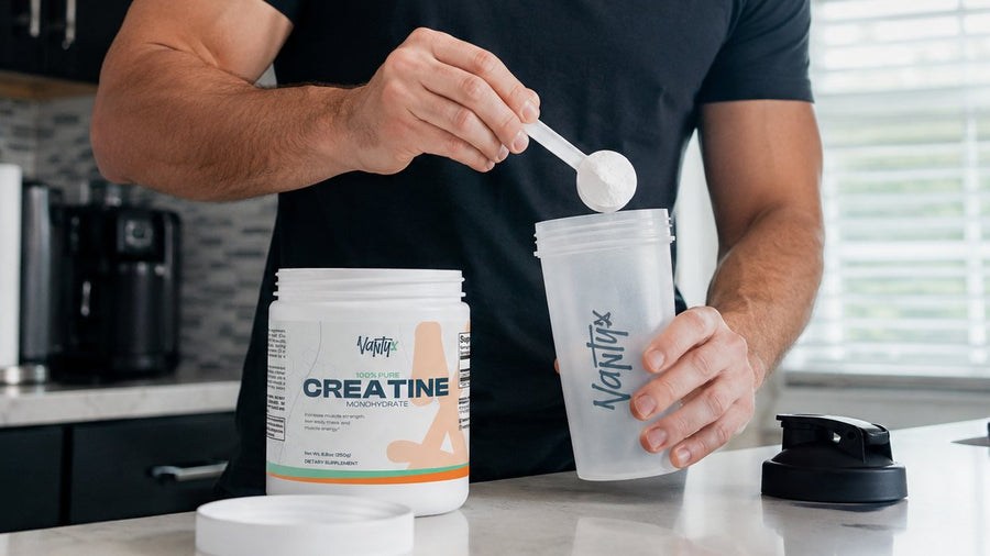 Scooping a 5 g serving of creatine monohydrate powder from an open tub into a shaker on a kitchen counter