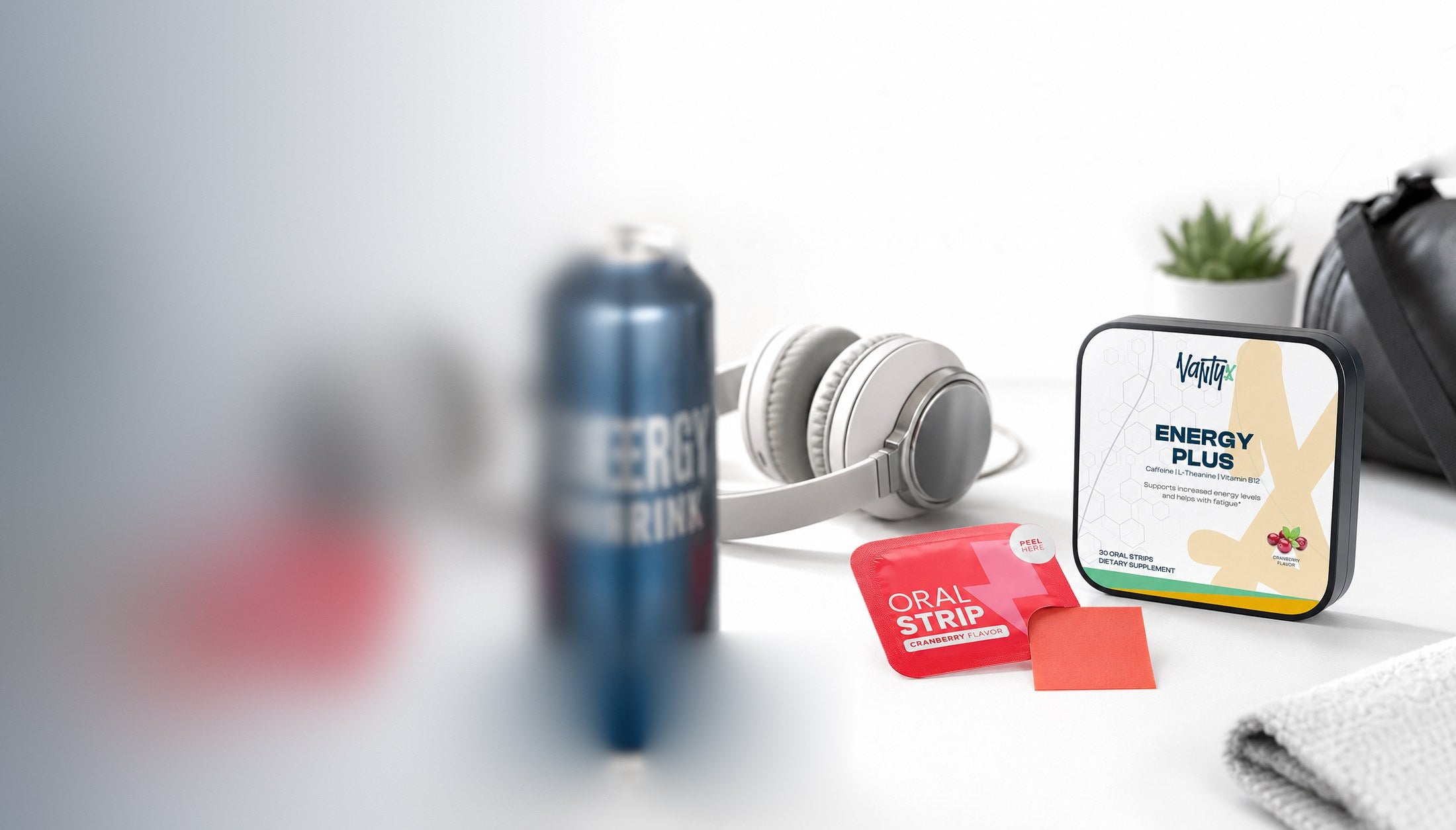 Vantyx Energy Plus tin and a cranberry oral strip pouch on a white desk beside headphones and an energy drink can